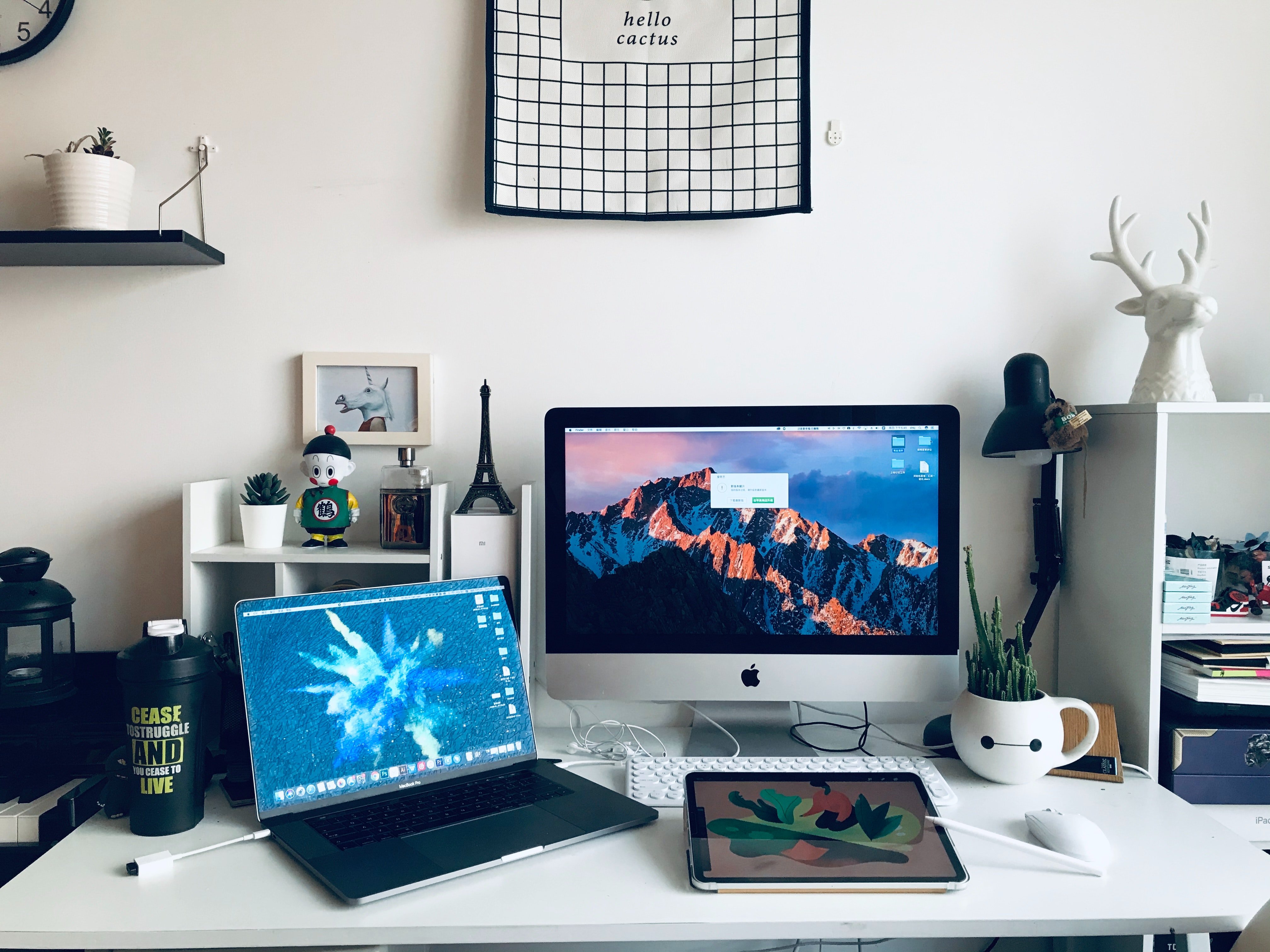 Büro-Setup mit einem PC-Bildschirm, einem Laptop und kleinen Regalen mit Deko und Pflanzen.